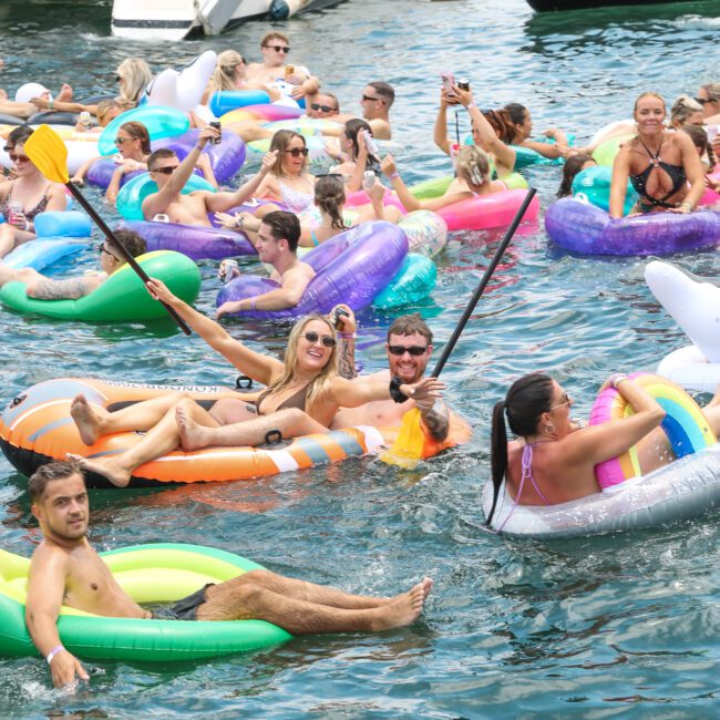 A lively group of people enjoying a sunny day on colorful inflatables in the water, with a sailboat in the background. Some are holding selfie sticks to capture the moment.