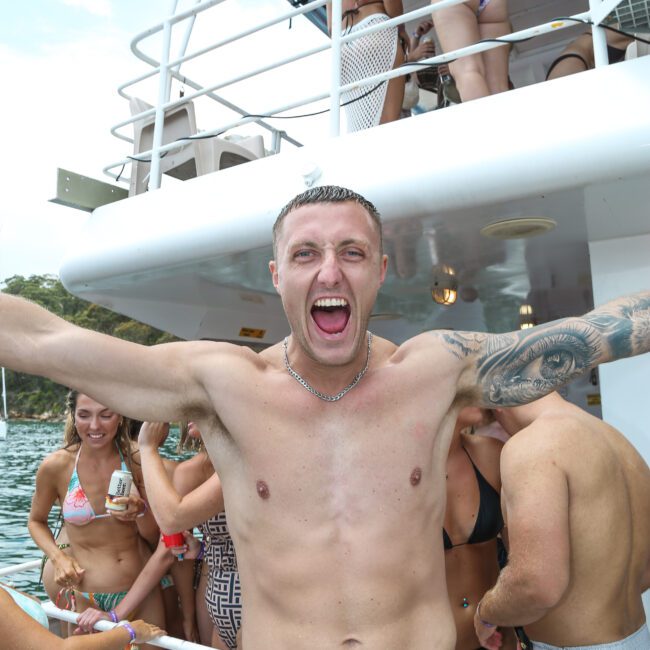 A man with tattoos, wearing black swim trunks, stands with arms wide open and mouth open on a boat with other people in swimwear. The scene is festive, with a backdrop of water and trees.