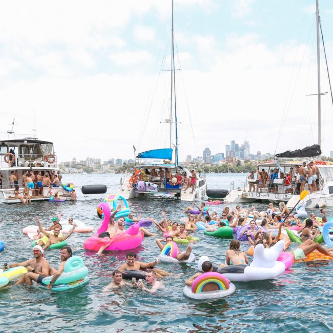 A group of people enjoying a sunny day on a harbor, floating on colorful inflatable toys in the water. Two boats are anchored nearby, and people are socializing. The city skyline is visible in the background under a partly cloudy sky.