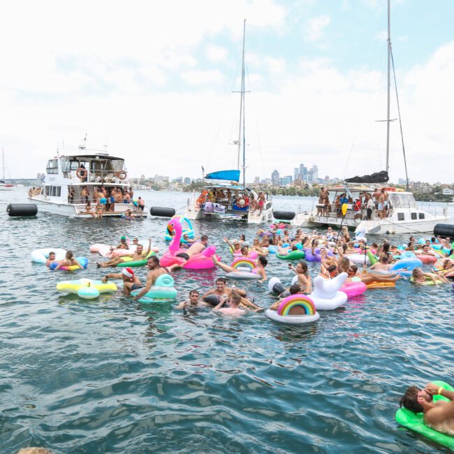 A large group of people enjoying a fun day on the water with colorful inflatable floats. Sailboats are anchored nearby, and the skyline is visible in the background. The scene is lively and festive under a bright sky.