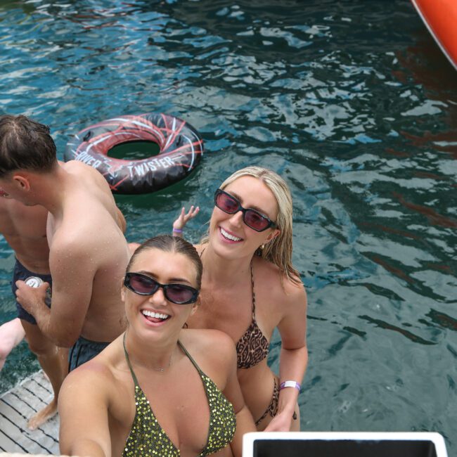 Two smiling people in swimsuits stand at the edge of a floating platform, with water and inflatable toys behind them. They appear to be enjoying a sunny day.