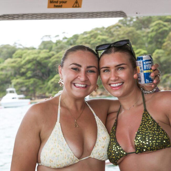 Two smiling women in swimsuits stand side by side on a boat. One holds a can of drink. There's a "Mind your head" sign above them, and a scenic view of trees and water in the background.