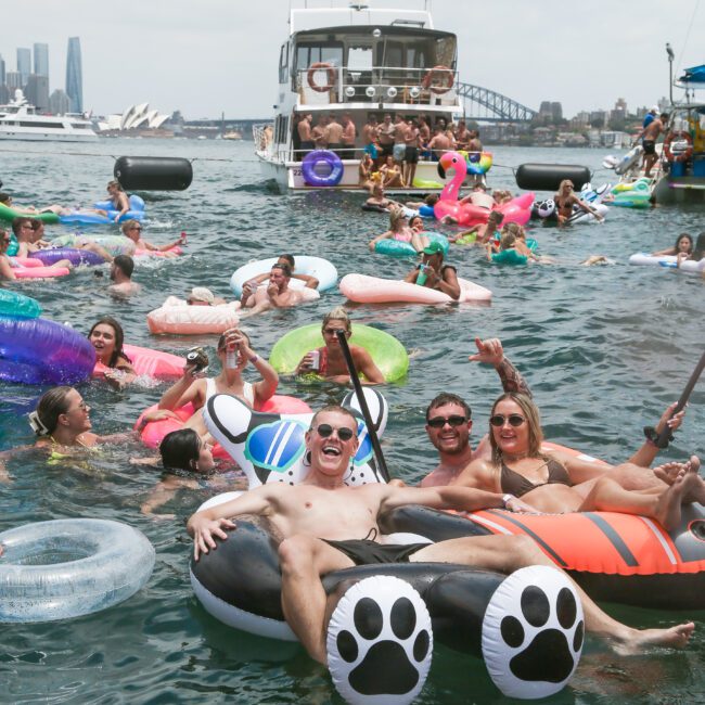 A group of people enjoying a sunny day on the water, floating on inflatable rafts shaped like animals. They're cheering and relaxing near boats, with a cityscape and a bridge visible in the background.