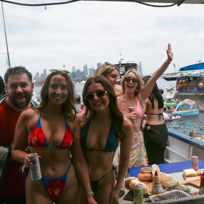 A group of people in swimsuits enjoy a boat party. Two women in bikinis pose in the foreground, smiling. Other individuals are behind them, with some waving. The city skyline and boats are visible in the background. Food and drinks are on the table.