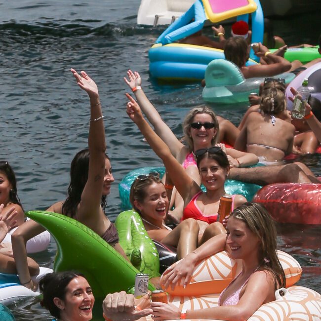 A group of people relax on colorful inflatable pool floats, holding drinks and enjoying the water. Some are laughing and waving, with a boat partially visible in the background. The atmosphere is festive and summery.