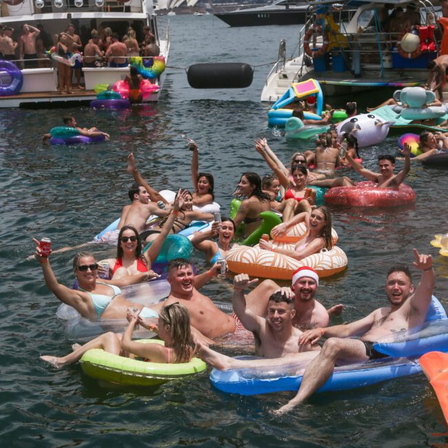 A group of people in colorful inflatable rafts enjoying a sunny day on the water, surrounded by boats. They are smiling, waving, and posing for the camera, suggesting a lively, festive atmosphere.