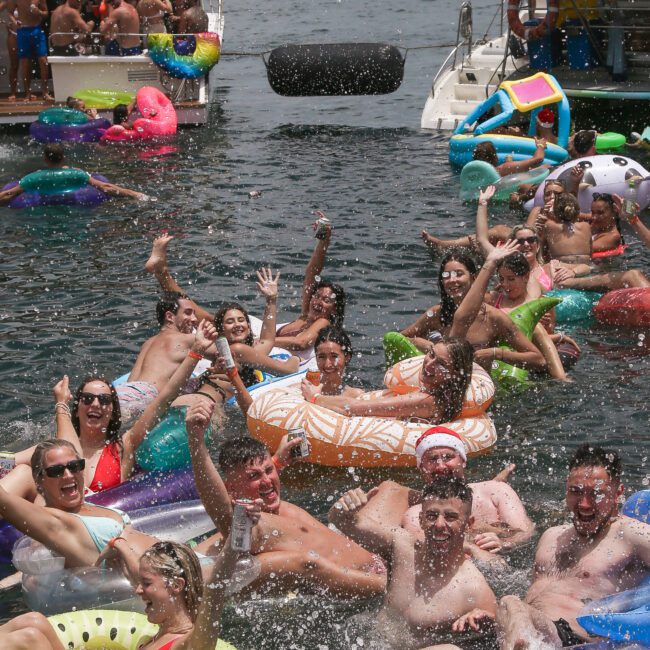 A group of people enjoying a sunny day on an urban waterway, lounging on colorful inflatable rafts and floaties. Boats are anchored nearby, and the iconic Sydney Opera House is visible in the background. Everyone appears to be having fun.