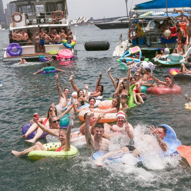 A group of people are having fun at a floating party on a river. They're wearing swimsuits and using inflatable floats. Boats are nearby, and the atmosphere is lively with everyone enjoying the water under a cloudy sky.