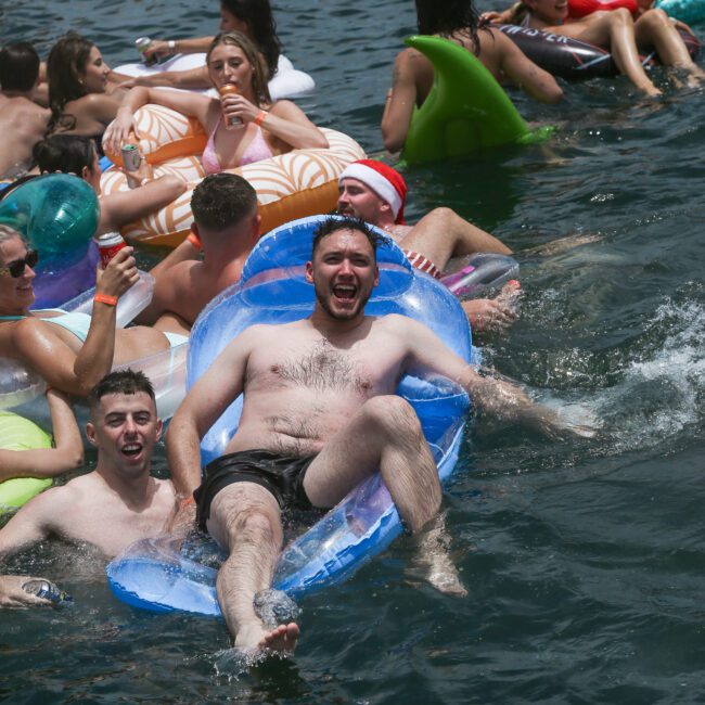 A group of people enjoying a sunny day in a large body of water. Some are on inflatable floats, including a person on a blue float and others on colorful ones. They appear to be having fun and socializing.