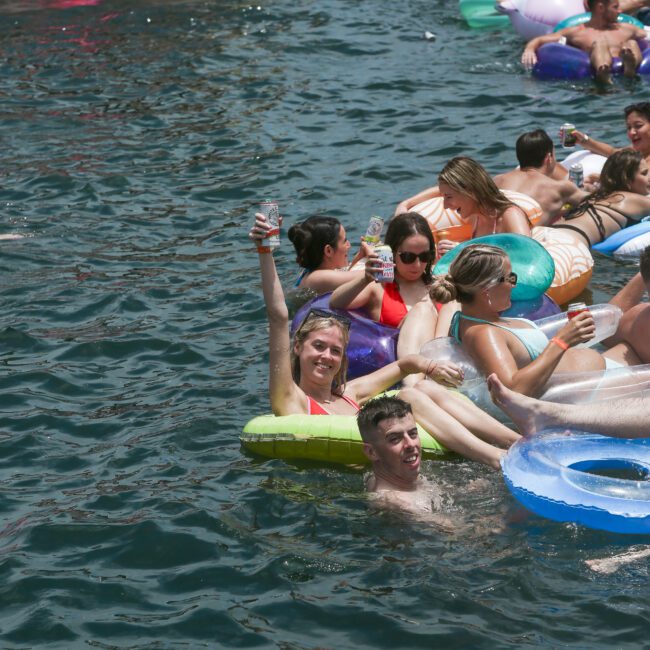 People relaxing on colorful inflatable floats in a body of water near a boat, enjoying a sunny day. One person holds up a drink, smiling. The scene is lively and filled with fun.