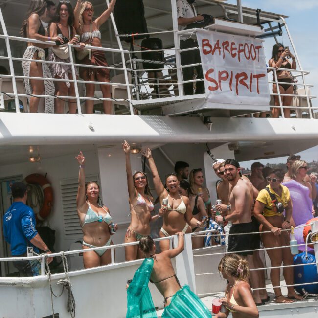 A group of people in swimwear are enjoying a party on a boat. Some are dancing and holding drinks, while others watch from the upper deck. A sign reads "Barefoot Spirit." The atmosphere is lively and festive.