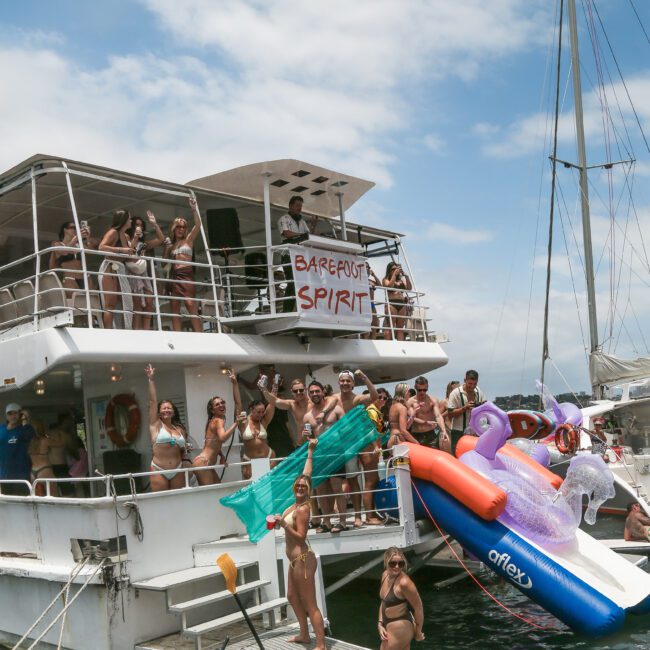 A lively group of people in swimsuits are partying on a boat labeled "Barefoot Spirit." Some are standing on a lower deck, with inflatables like a unicorn and kayak around them, enjoying the sunny weather and festive atmosphere.