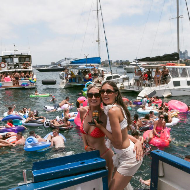 Two women smiling on a boat with a large group of people swimming and lounging on inflatables in the water. Boats are anchored nearby under a partly cloudy sky. The mood is lively and festive.