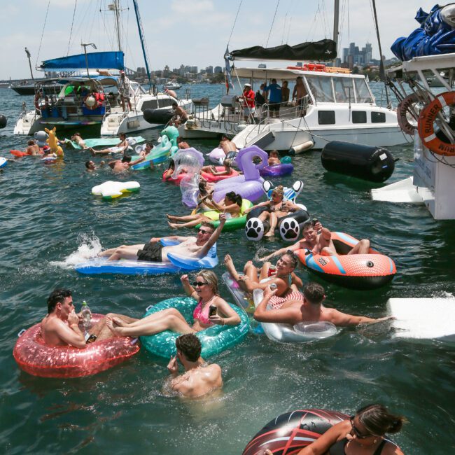 A lively scene of people enjoying a sunny day in a harbor, floating on colorful inflatables near several boats. The water is bustling with activity, featuring various inflatable shapes, including animals and tubes.