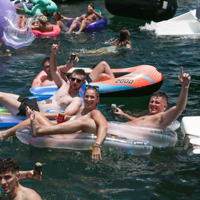 A group of people relaxing on colorful inflatable floats in the water near a boat. They're smiling, holding drinks, and enjoying a sunny day. More floats and a boat with people are visible in the background.