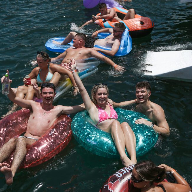 A group of people enjoy a sunny day, lounging on colorful inflatables in the water near a boat. They are smiling and appear relaxed, with some holding drinks. The scene conveys a festive, summer atmosphere.