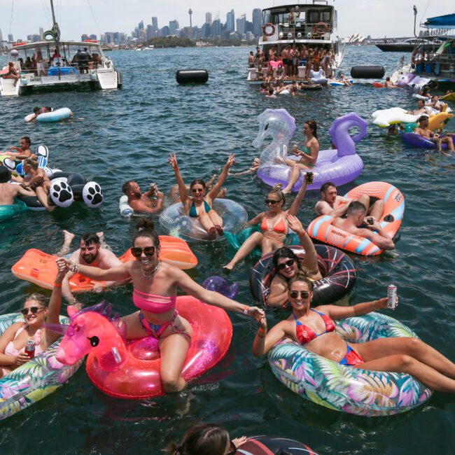 A lively group of people relax on colorful inflatable floats in a large body of water. Boats are anchored nearby, and the skyline is visible in the background. The scene captures a summer party atmosphere.