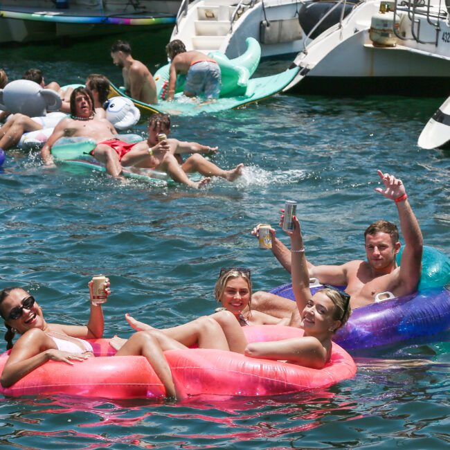 A group of people are enjoying a sunny day on a lake, lounging on colorful inflatable floats. Boats are docked nearby, and the scene is lively with people smiling, holding drinks, and relaxing in the water.