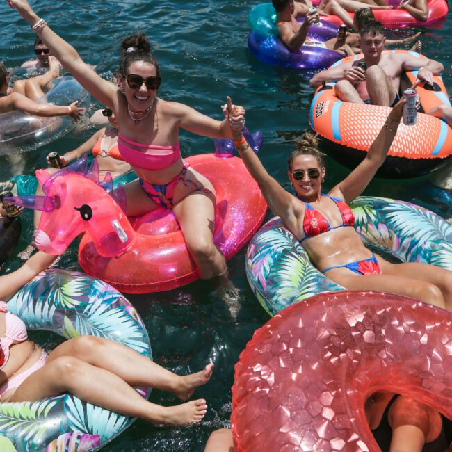 A group of people enjoying a sunny day on a lake, floating on colorful inflatable tubes. Two women are smiling and raising their arms, one on a pink flamingo float and the other on a leafy-patterned float. The atmosphere is lively and festive.