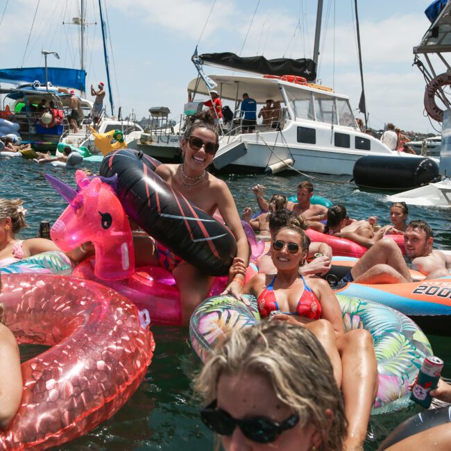 A group of people enjoying a sunny day on inflatable floats in the water, surrounded by boats. One person sits on a pink unicorn float, smiling. Others lounge on various colorful floats, and sailboats are anchored in the background.