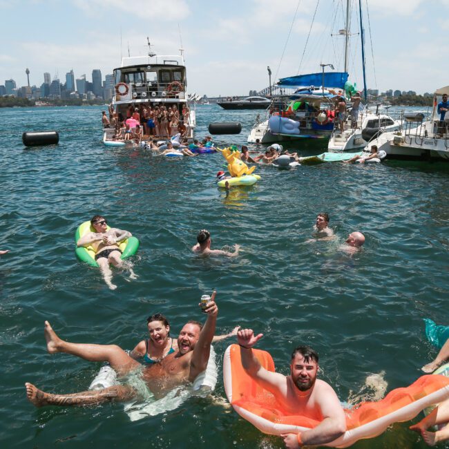 A lively group enjoys a sunny day on the water, lounging on colorful inflatable floats near boats. The city skyline is visible in the background, adding to the festive atmosphere.