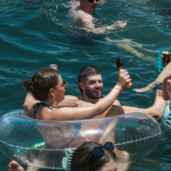 A group of people enjoying a sunny day in the water. A man and woman are seated on a float, with the man taking a selfie. Others swim and relax nearby, surrounded by inflatables in a lively, fun atmosphere.