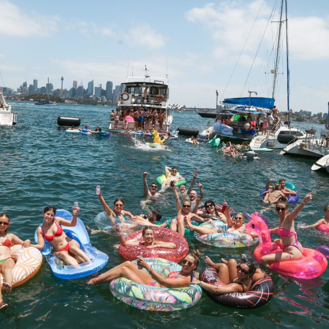 A lively scene with people relaxing on inflatable floats in a harbor. Several boats surround them as a city skyline and blue sky form the backdrop. The atmosphere is festive and joyful, with everyone enjoying a sunny day on the water.