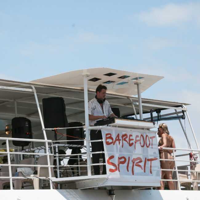 A DJ is performing on the deck of a boat decorated with a banner that reads "Barefoot Spirit." People in swimwear are enjoying the sunny day on the boat.