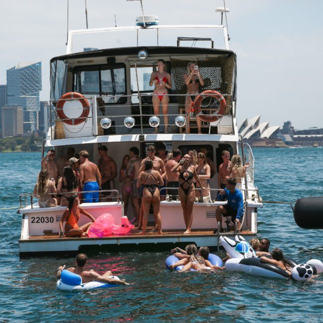 A group of people on a yacht partying, with several more in the water nearby on inflatable floats. The skyline of Sydney, including the Opera House, is visible in the background under a clear blue sky.