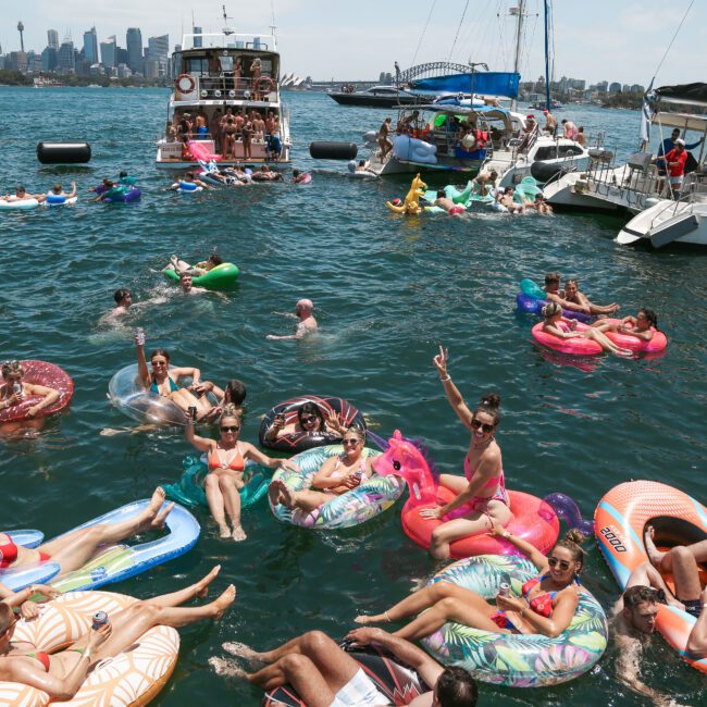 A group of people relax on colorful inflatable tubes and floaties in the water, surrounded by boats. The scene is lively under a clear sky, with a city skyline visible in the background.