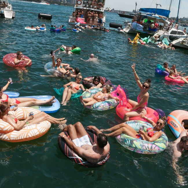 People relax on colorful inflatable tubes in a sunny harbor, with boats and a city skyline in the background. The lively scene includes various floats, such as swans and donuts. A person gives a peace sign, enjoying the vibrant atmosphere.