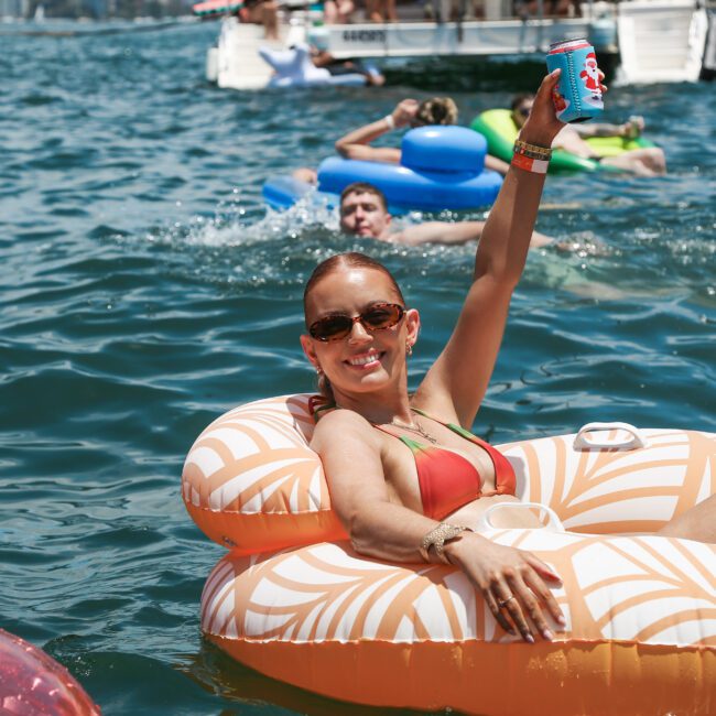 A person in a red swimsuit relaxes on a patterned inflatable float in a lake, holding up a drink. Other people are floating in the water and a boat is visible in the background on a sunny day.