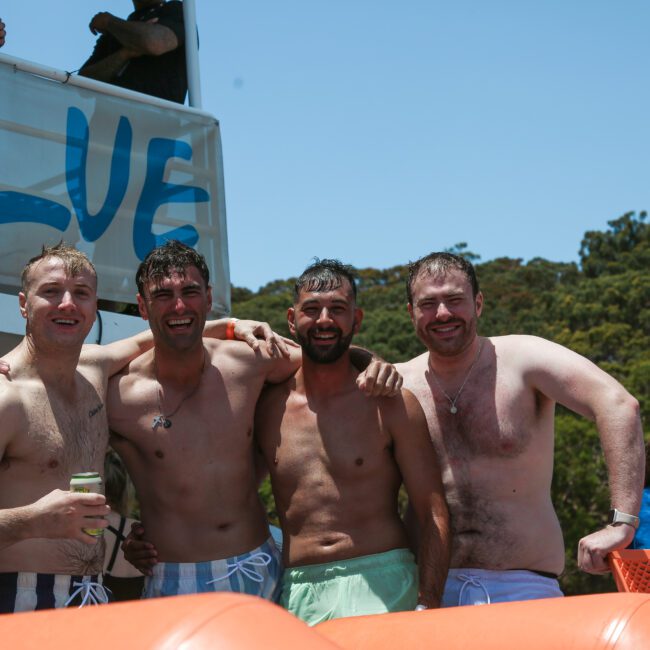 Four men in swim trunks stand close together on a boat, smiling at the camera. Behind them are trees and a sunny sky. Other people are visible in the background, and part of the boat with the word "BLUE" is in view.