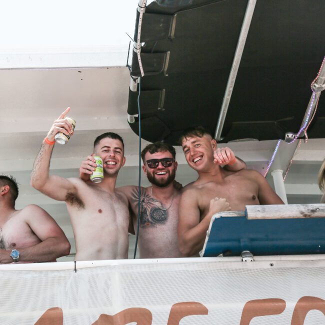 A group of four shirtless people, three men and one woman, are on a boat. Three men are smiling at the camera, with one holding a drink. The woman is seated, wearing sunglasses, and also smiling. The boat has a black canopy overhead.