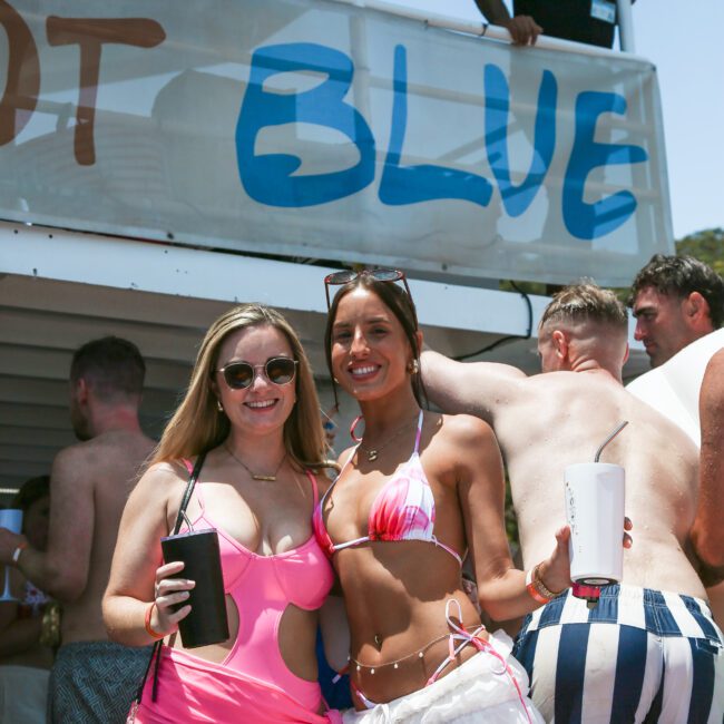 Two women smiling on a boat, wearing swimsuits and holding drinks. One is in a pink outfit with sunglasses, the other in a bikini. People are gathered on the deck above. The partial sign reads "BLUE." A sunny day with clear skies.