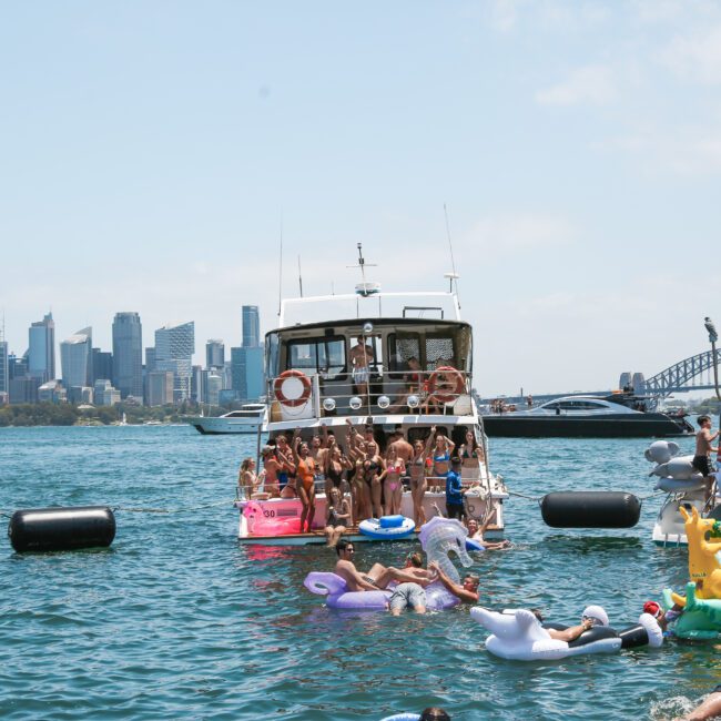 A group of people enjoying a sunny day on a boat in Sydney Harbour. The water is filled with colorful inflatable floats shaped like a unicorn, duck, and swan. The city skyline and Sydney Harbour Bridge are visible in the background.