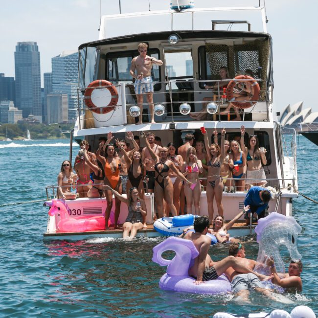 A group of people enjoying a boat party on the water with the Sydney skyline in the background. They are in swimsuits, with some on the boat and others on inflatable pool floats in the water. The atmosphere is festive and lively.