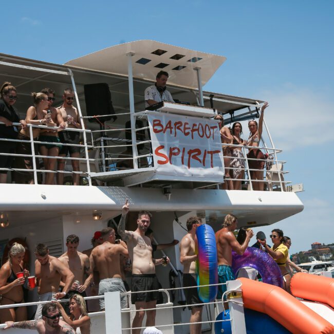 A festive group of people enjoying a party on a boat labeled "Barefoot Spirit." The scene includes individuals on the upper deck and in a swimming area with inflatable slides. Clear blue sky and coastal scenery are visible in the background.