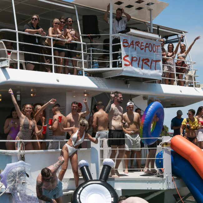 A group of people enjoying a sunny day on a boat called "Barefoot Spirit." Some are on the deck waving, while others are at the lower level with inflatable tubes and a slide. The sky is clear and blue.