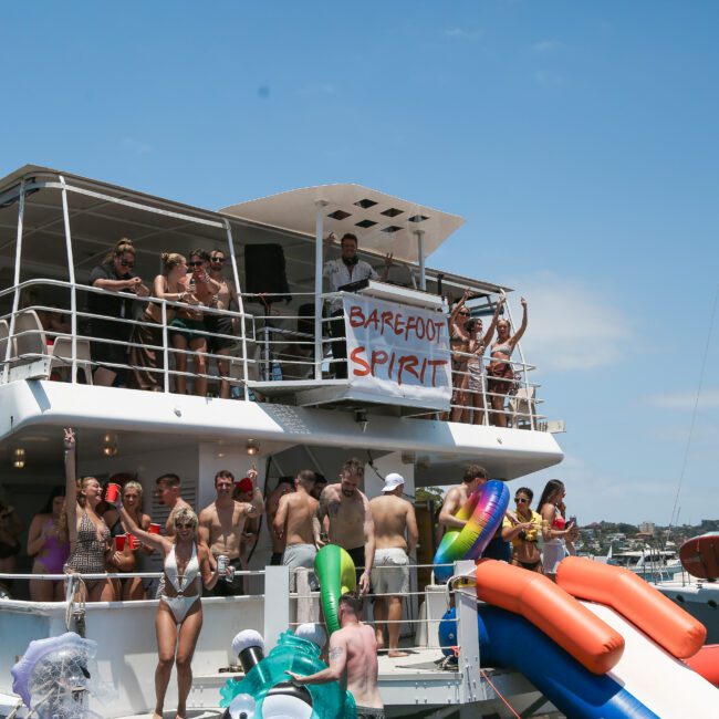 A group of people enjoying a party on a two-level yacht named "Barefoot Spirit." Some participants are dancing, and others are lounging by an inflatable water slide. The sky is clear and blue, suggesting a sunny day.