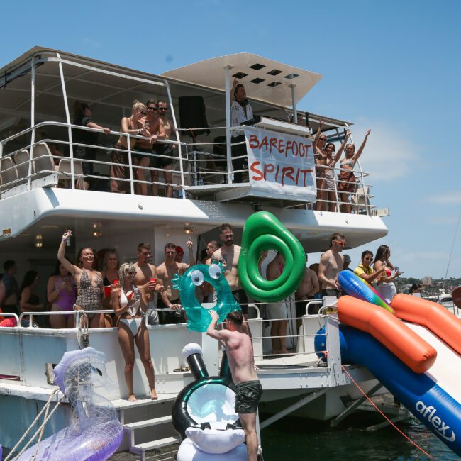 A group of people enjoying a party on a boat with a sign reading "Barefoot Spirit." The scene features many wearing swimsuits, and large inflatable floats shaped like unicorns and other creatures. Bright and festive atmosphere under clear skies.