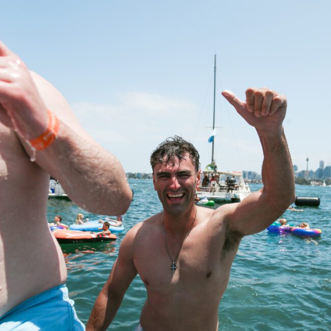 A man in swim trunks smiles and gives a thumbs up while standing by the water. Other people and inflatable floats are in the background, with boats and a clear sky visible.