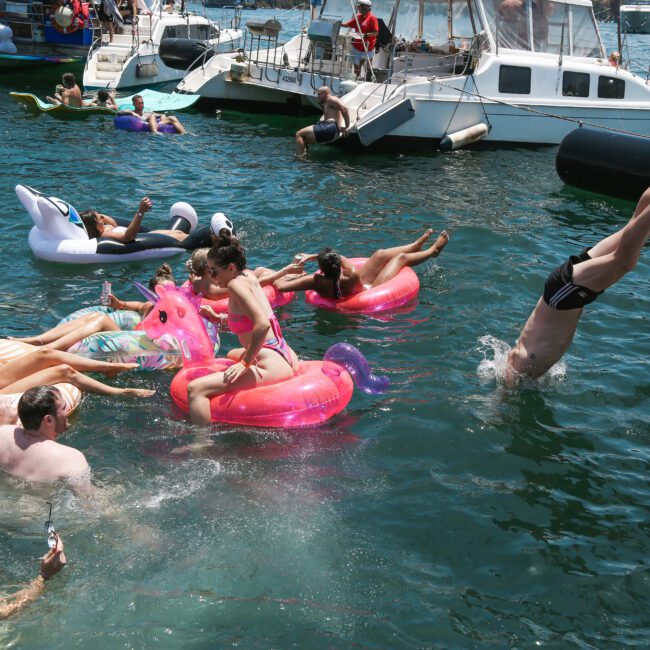 People enjoy the water near boats, lounging on colorful inflatable floats shaped like unicorns and swans. One person dives into the water from a boat, and several others relax and chat nearby.