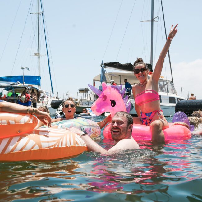 A group of people enjoying a sunny day on the water, relaxing on inflatable pool floats, including a pink unicorn. Sailboats are anchored in the background, and everyone appears to be having fun.