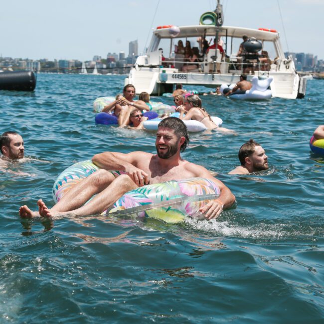 A group of people enjoying a sunny day in the water with colorful inflatables. A boat is anchored nearby, and a city skyline is visible in the background. Everyone appears relaxed and happy.