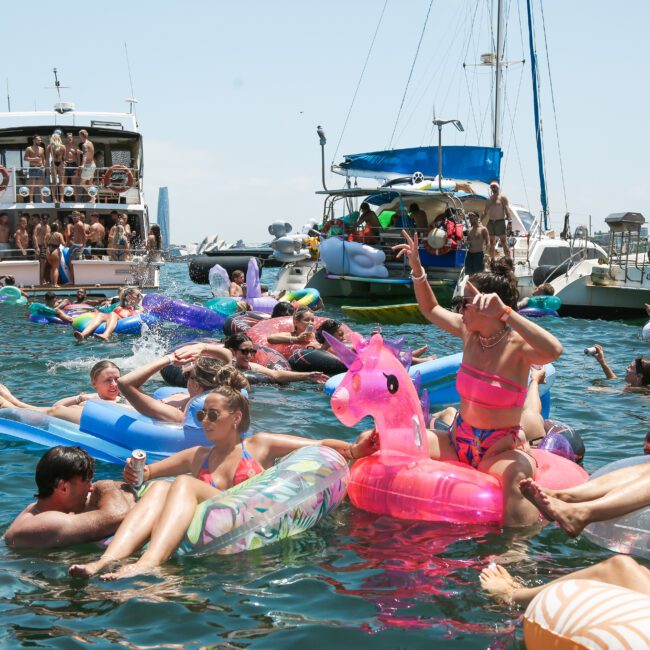 A group of people enjoying a sunny day on the water, lounging on colorful inflatable floats, including unicorns and flamingos. Boats are anchored nearby, and the city skyline is visible in the background.