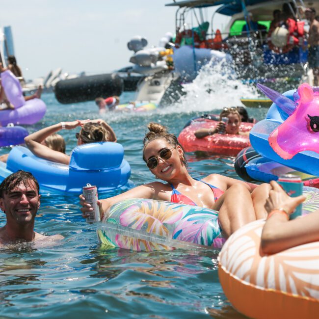 A group of people enjoying a boat party on a sunny day. They are relaxing on colorful inflatable floats, including a unicorn-shaped one, in the water. Boats are visible in the background. The atmosphere is lively and festive.