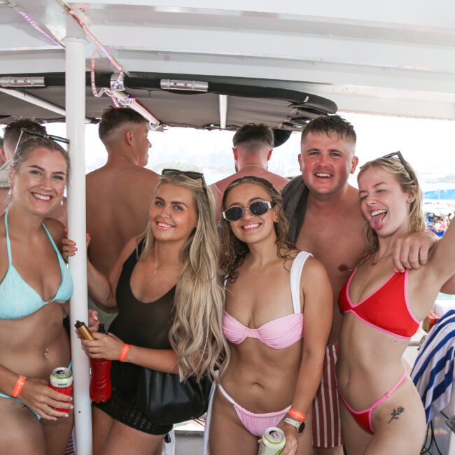 A group of people in swimwear are smiling and posing on a boat. They appear to be enjoying a sunny day on the water, with drinks in hand. The sea and other boats can be seen in the background.