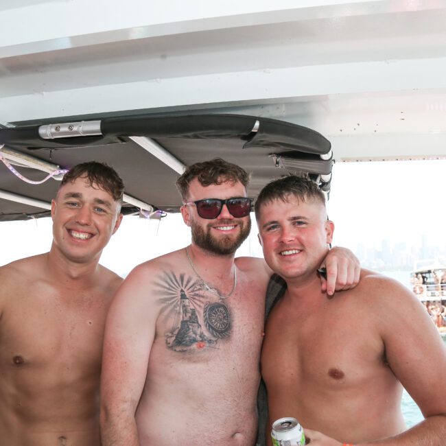 Three shirtless men smile at the camera on a boat. The man in the center wears red sunglasses and shows a chest tattoo. The background features a city skyline and several people swimming and enjoying the water.