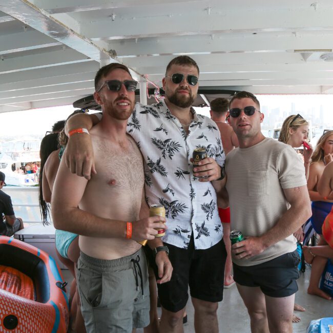 Three men posing together on a boat. They're wearing casual summer clothes and sunglasses, holding drinks. Other people are in the background, enjoying the sunny day on the water. An orange inflatable is on the boat's deck.